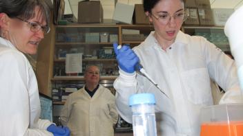 Two women and a man in white coats at a lab bench; one woman pipetting